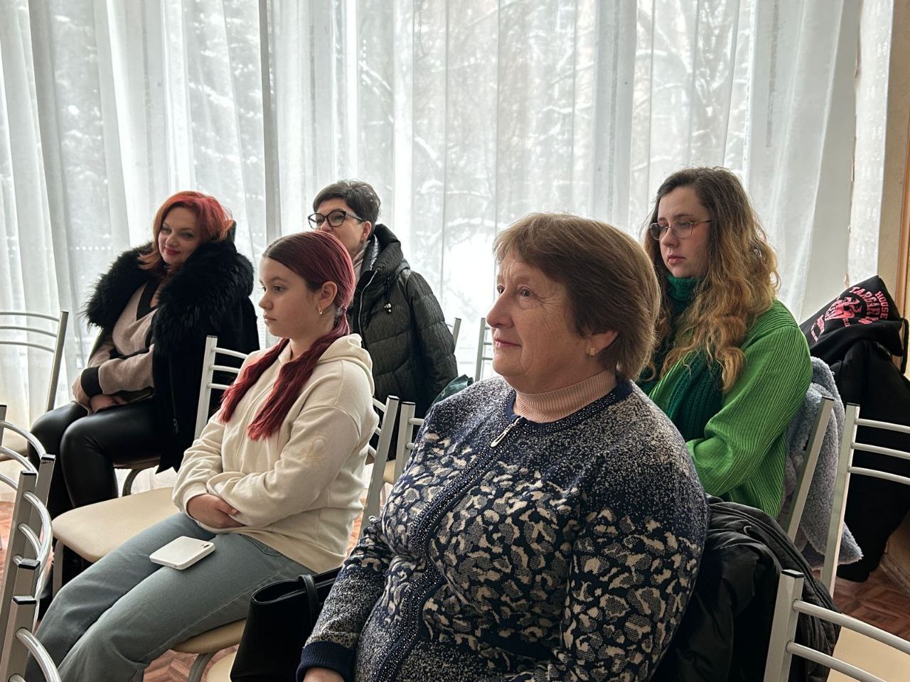 Група жінок різного віку сидить на стільцях у світлій залі біля вікна та уважно слухає виступ. Жінки одягнені в теплий повсякденний одяг: светри, жилети та куртки.
