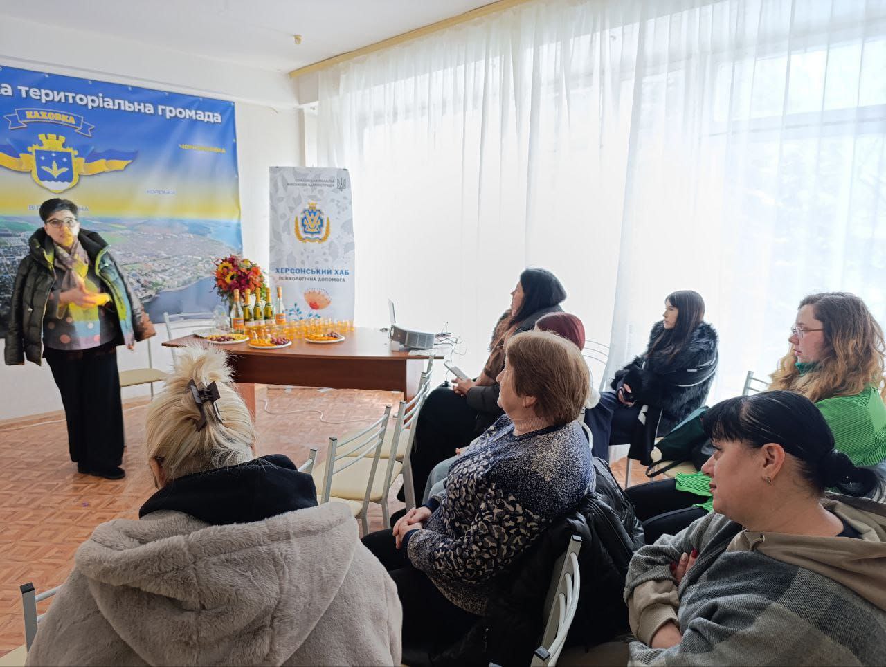 Жінка проводить презентацію для невеликої аудиторії у світлій залі. У центрі стоїть стіл із частуваннями, а на стінах розміщено банер територіальної громади та інформаційний стенд психологічної допомоги «Херсонський хаб».