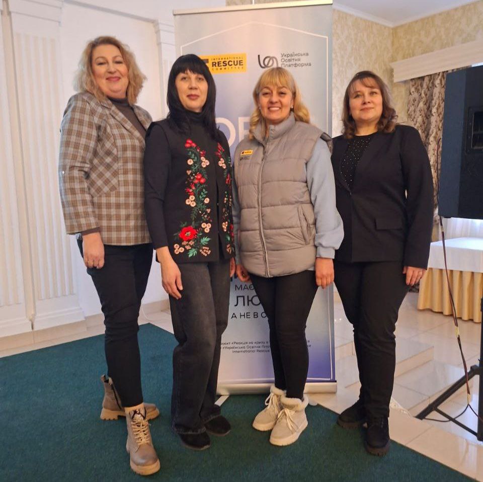 Чотири жінки стоять поруч і посміхаються на фоні інформаційного банера в офіційному приміщенні. Позаду видно білі колони та частину музичного обладнання.