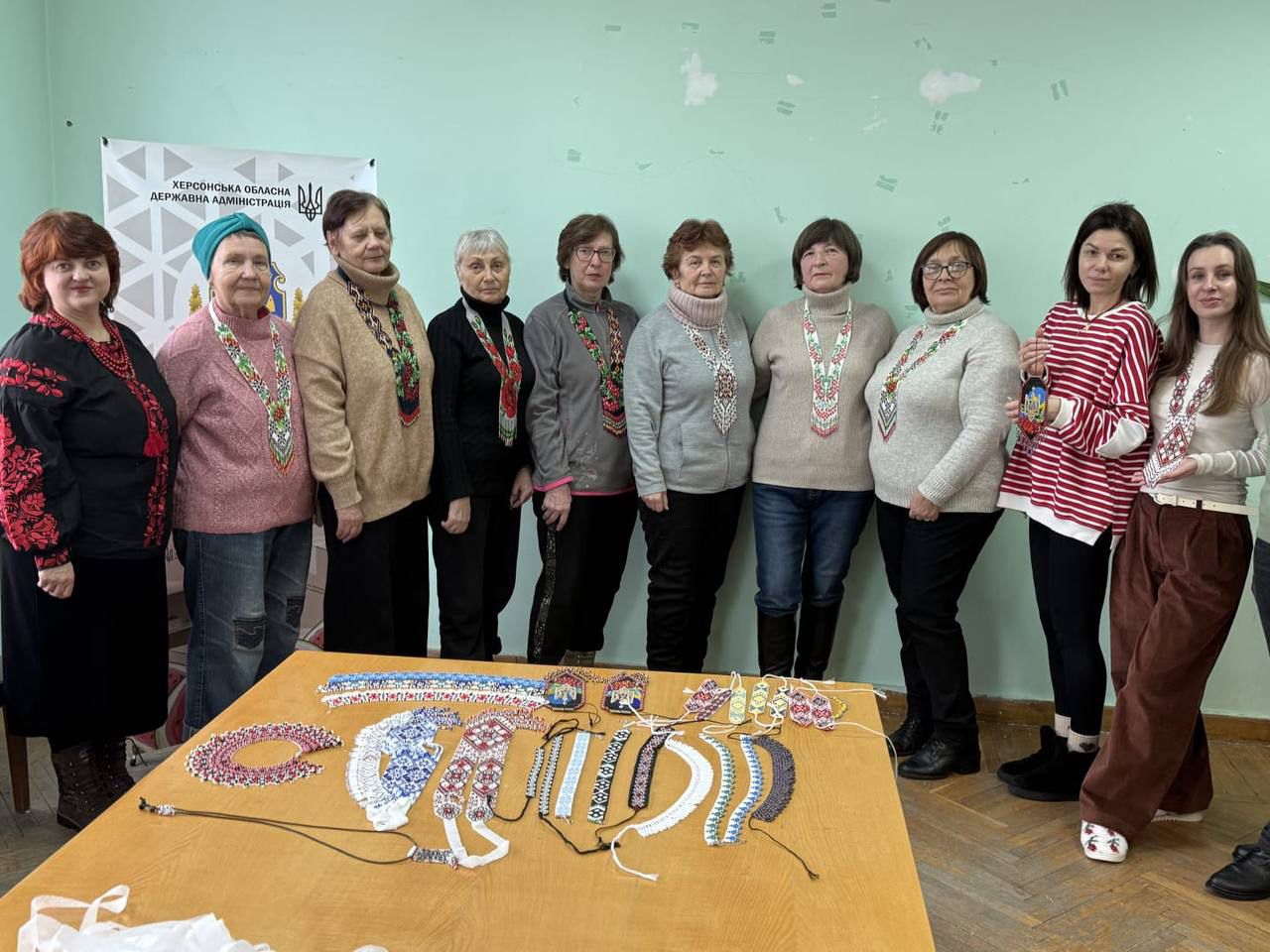 Група жінок стоїть біля столу, на якому розкладені різнокольорові прикраси з бісеру ручної роботи. На фоні видно банер Херсонської обласної державної адміністрації, а жінки одягнені у вишиванки або мають прикраси в етнічному стилі.