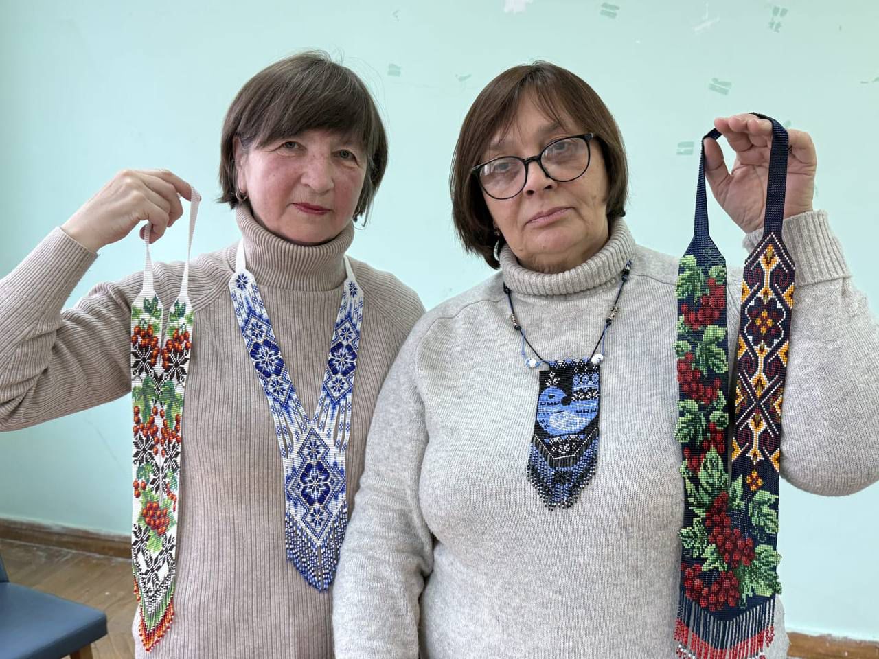 Дві жінки демонструють виготовлені вручну прикраси з бісеру (гердани) з традиційними українськими орнаментами. На одній прикрасі зображено калину, на інших — геометричні візерунки у синьо-білих та червоно-чорних кольорах.
