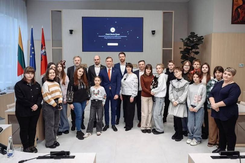 Групове фото великої компанії дорослих та підлітків у сучасному залі для офіційних зустрічей на фоні прапорів та екрана з технічним написом. Деякі учасники вдягнені у вишиванки.