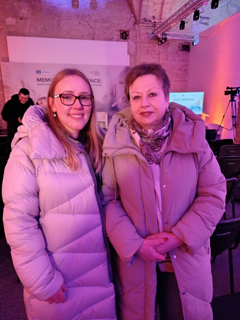 Дві жінки у теплих світлих куртках стоять поруч у великій залі з рожевим освітленням. На задньому плані видно банер конференції ЮНЕСКО, ряди стільців та професійну відеокамеру на штативі.