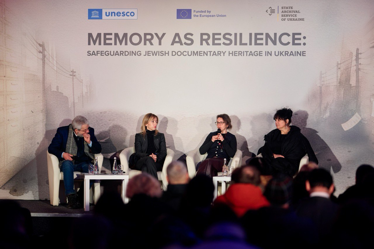 Чотири спікери беруть участь у дискусії на фоні великого банера конференції ЮНЕСКО «Memory as Resilience: Safeguarding Jewish Documentary Heritage in Ukraine». У залі присутня аудиторія, яку видно зі спини на передньому плані.