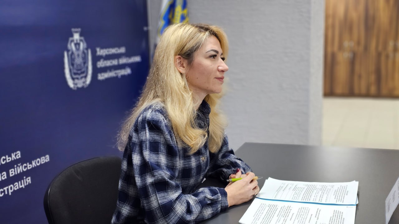 Жінка у синій картатій сорочці сидить за столом під час офіційного заходу. Вона тримає ручку, перед нею лежать документи. Позаду неї — синій банер з гербом та написом «Херсонська обласна військова адміністрація».