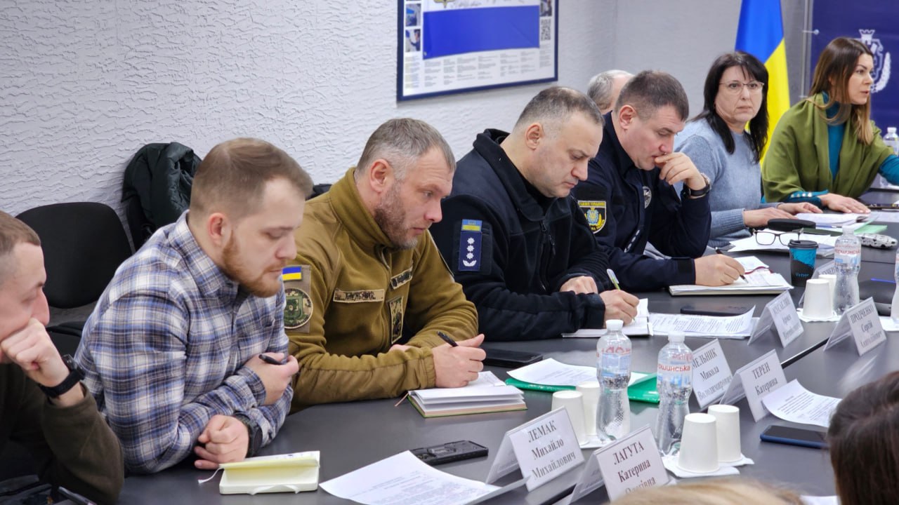 Ряд учасників наради за робочим столом. Чоловіки та жінки у цивільному одязі та військовій формі роблять записи у блокнотах. На столі стоять пляшки з водою та таблички з іменами. На задньому плані видно частину прапора України.