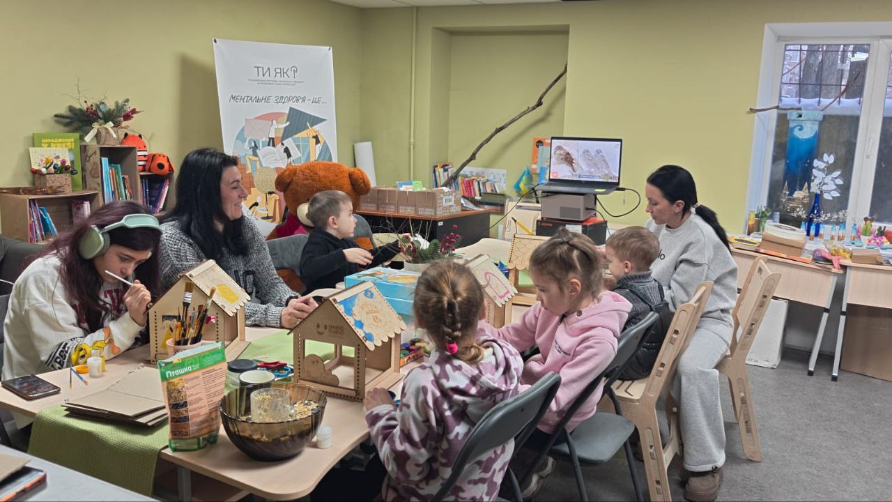 Дорослі та діти сидять за великим столом у творчій майстерні, розфарбовуючи дерев'яні годівнички для птахів. На фоні видно плакат програми ментального здоров'я та вікно.