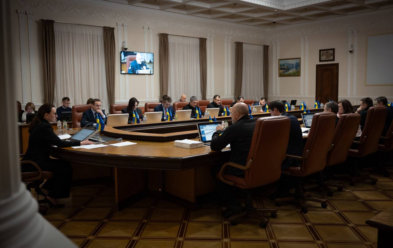 Засідання в офіційній залі за великим круглим столом. Учасники працюють із ноутбуками та документами, на стіні встановлено монітор, на якому транслюється відеозв’язок. Приміщення оздоблене класичними колонами, високими вікнами з гардинами та прапорцями на столах.