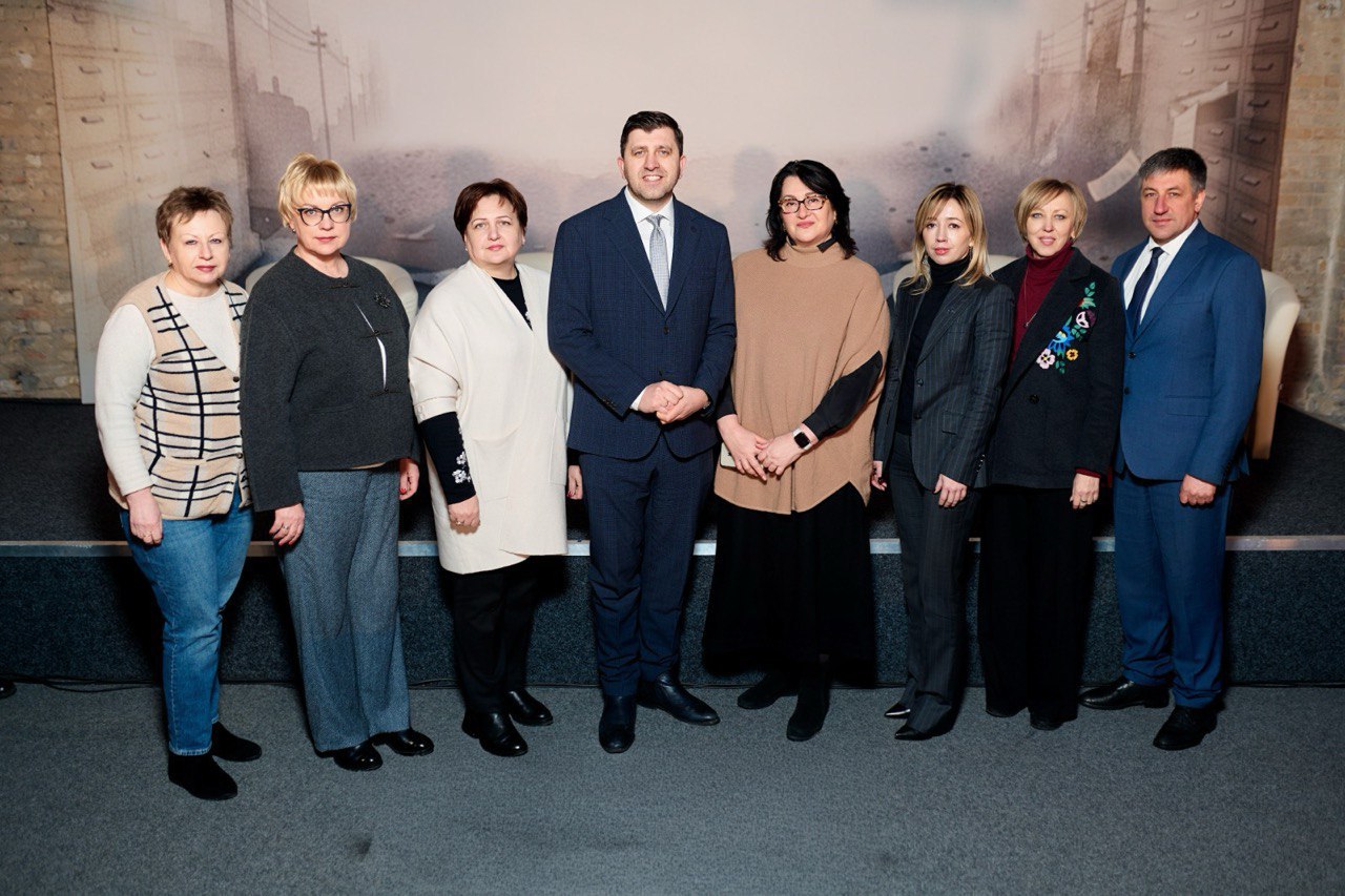 Група офіційно вбраних людей — двоє чоловіків у костюмах та п'ятеро жінок у діловому одязі — стоять у ряд на сцені під час офіційного заходу на фоні тематичного банера.