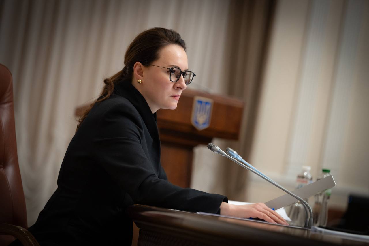 Жінка в окулярах та офіційному костюмі сидить за столом у залі засідань. Вона зосереджено дивиться перед собою, тримаючи руку на документах біля мікрофонів. На задньому плані видно дерев'яну трибуну з гербом України.