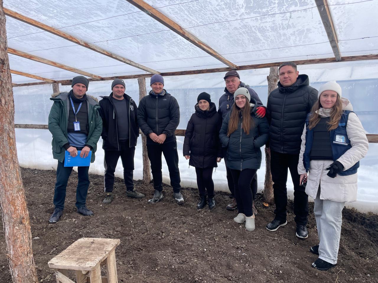 Група людей у зимовому одязі стоїть усередині великої теплиці з дерев'яним каркасом та плівковим покриттям. Учасники зустрічі позують на фоні підготовленого до посадки ґрунту. Деякі люди мають прикріплені бейджі, а чоловік ліворуч тримає синю папку з документами.