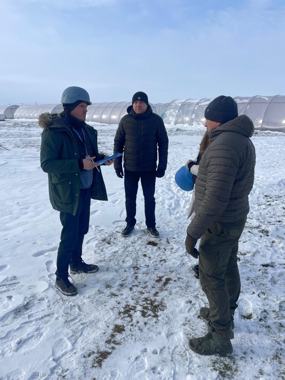 Троє чоловіків у зимовому одязі розмовляють на засніженому полі поруч із довгими тепличними конструкціями. Один із них одягнений у зелену куртку та захисний шолом, тримаючи в руках папку з документами. Інші учасники зустрічі одягнені в темні пуховики та шапки.