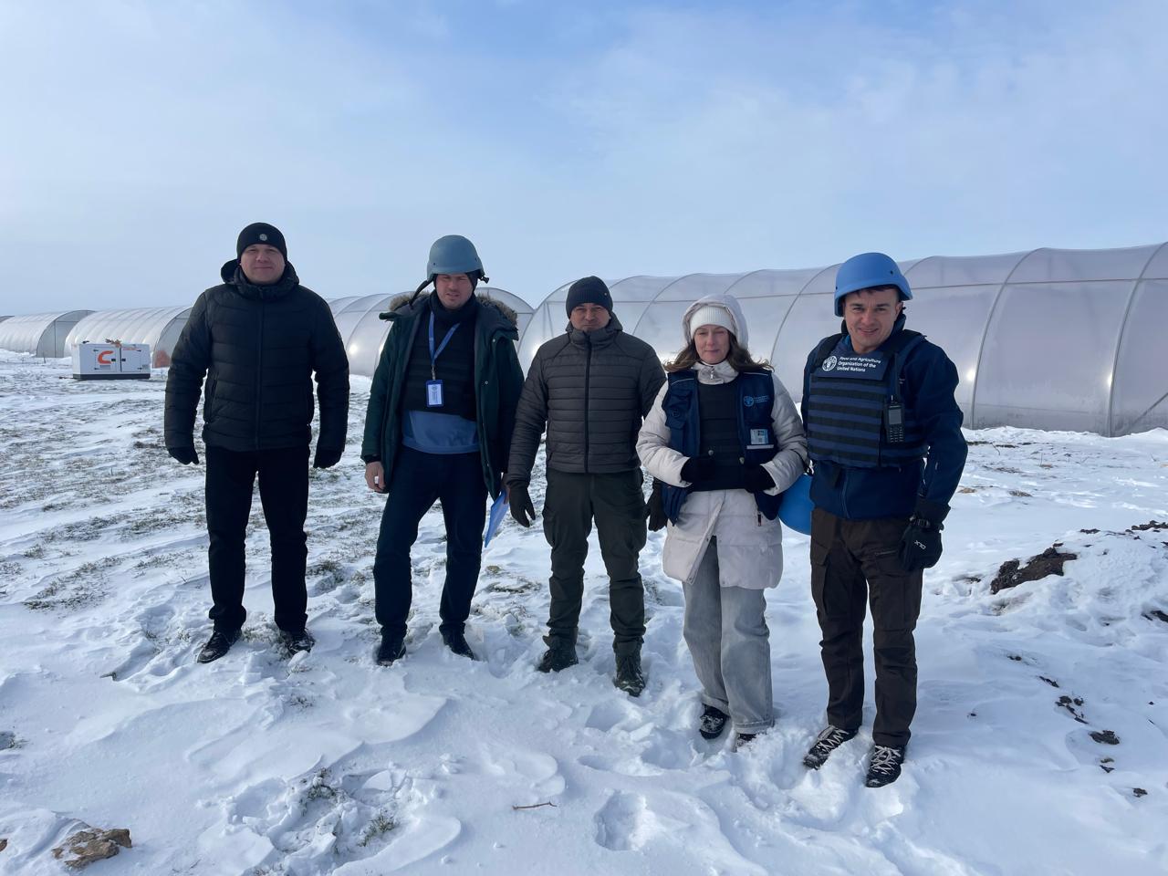 П’ятеро людей у зимовому одязі стоять на засніженому полі на фоні довгих напівсферичних теплиць. Двоє чоловіків одягнені в захисні шоломи та бронежилети з логотипом міжнародної організації, а жінка поруч — у світлий пуховик та жилет із бейджем. Усі учасники дивляться в камеру.