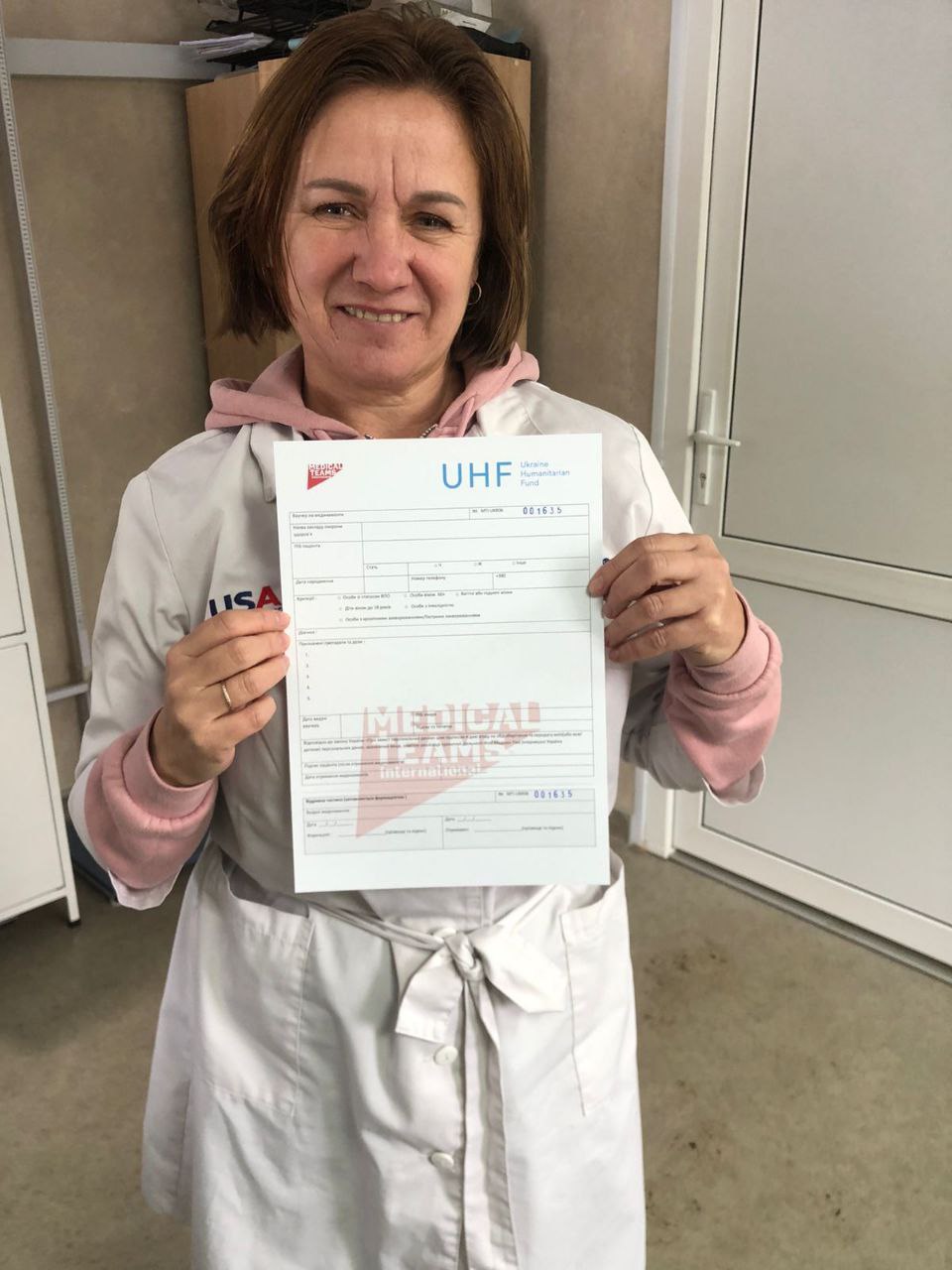 Жінка в білому медичному халаті тримає перед собою офіційний бланк документа з логотипами «Ukraine Humanitarian Fund» та «Medical Teams International».
