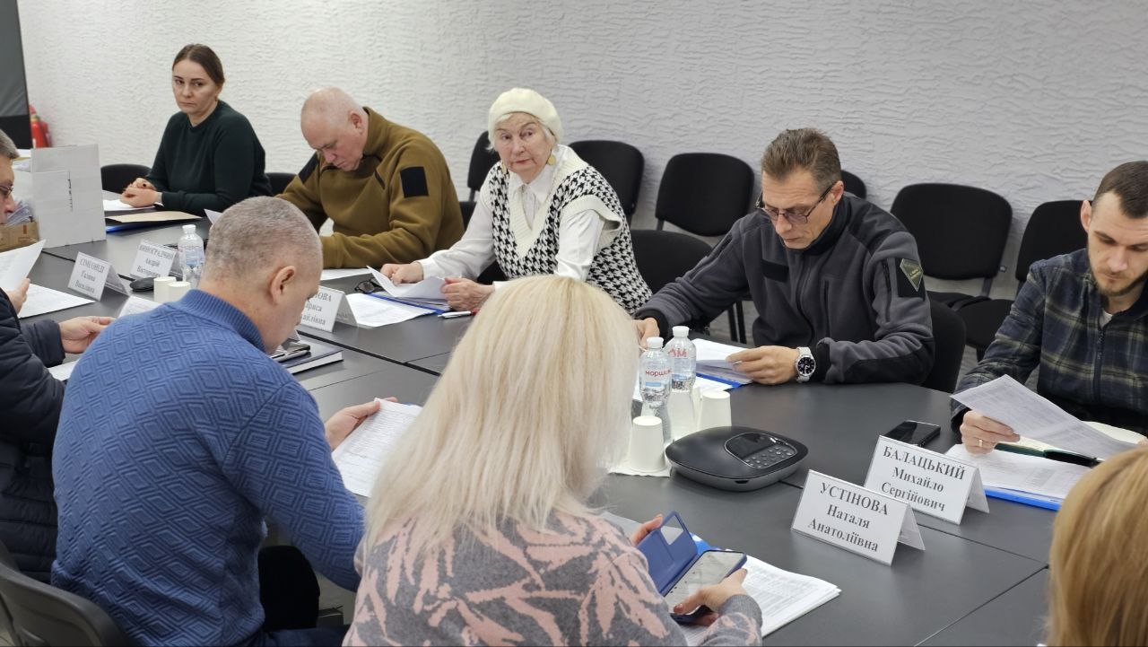 Група людей сидить за довгим робочим столом під час засідання. Учасники наради уважно вивчають паперові документи, деякі користуються смартфонами. На столі розміщені іменні таблички, пляшки з водою та офісна техніка. Атмосфера зосереджена, приміщення оформлене у світлих тонах.