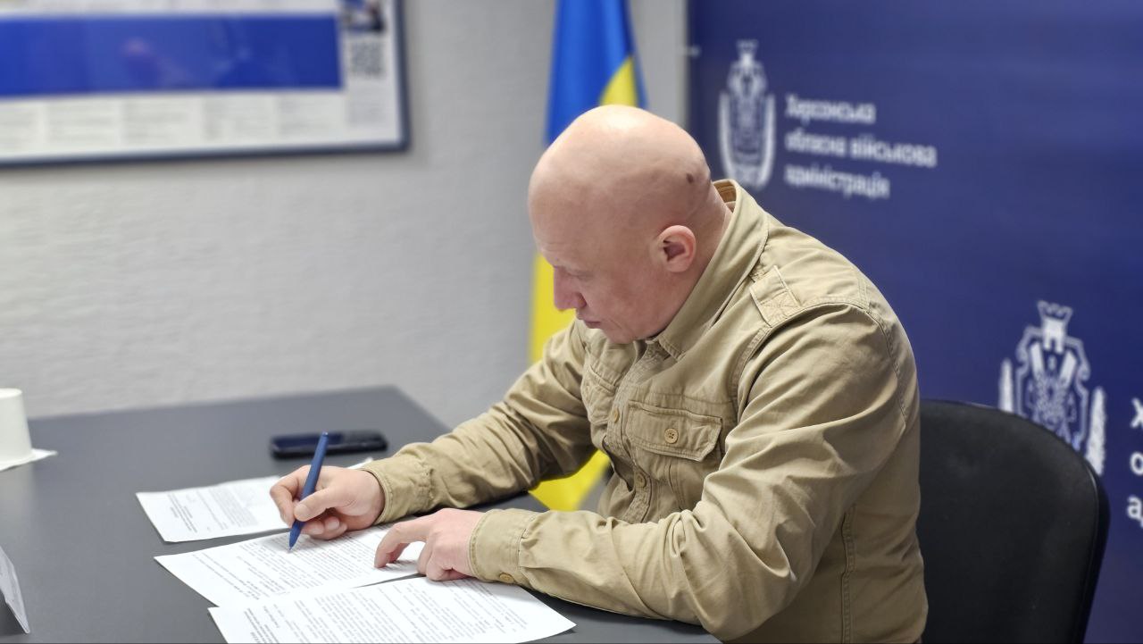Чоловік у тактичній сорочці пісочного кольору сидить за столом і підписує документи синьою ручкою. На задньому плані видно Державний Прапор України та частину синього банера з офіційною символікою Херсонської обласної військової адміністрації.
