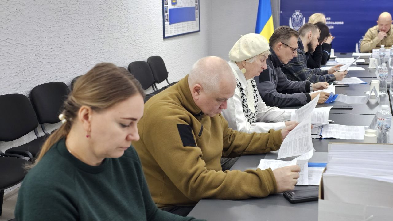 Учасники наради сидять за робочим столом в офіційному приміщенні та уважно вивчають багатосторінкові документи. На задньому плані видно Державний Прапор України. Люди одягнені у повсякденний та тактичний одяг, атмосфера робоча та зосереджена.