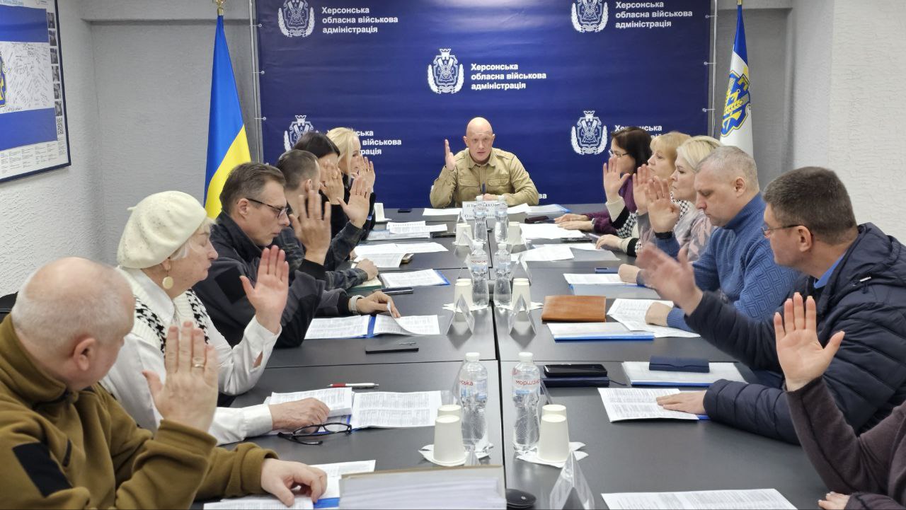Процес голосування під час робочої наради в офіційному приміщенні. Група людей за довгим столом одночасно піднімає руки. У центрі, на фоні синього банера «Херсонська обласна військова адміністрація», сидить чоловік у військовій формі. Учасники наради — чоловіки та жінки різного віку в цивільному та військовому одязі.