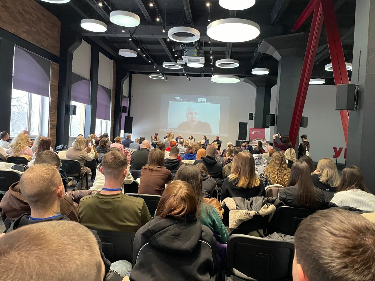 Велика аудиторія в сучасному стилі, заповнена людьми, які сидять спиною до камери та дивляться на екран. На великому центральному екрані транслюється відеовиступ чоловіка у форматі онлайн-конференції. Стеля приміщення оздоблена численними круглими плафонами, що створюють яскраве освітлення.