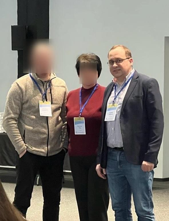 Троє учасників конференції стоять поруч у світлому приміщенні. На кожному з них одягнений бейдж на синій стрічці. Чоловік праворуч одягнений у синій піджак, світлу сорочку та джинси; обличчя інших двох учасників приховано.