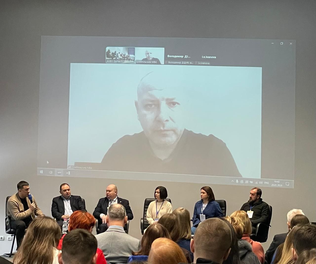 Панельна дискусія під час конференції. Шестеро спікерів сидять на сцені перед аудиторією та обговорюють робочі питання. Над ними на великому екрані транслюється відеовиступ іншого учасника у форматі онлайн-включення.