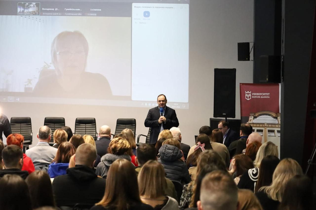 Виступ спікера на фоні великого екрана у залі, заповненій людьми. На екрані транслюється онлайн-включення іншої учасниці заходу. Праворуч розміщено банер із логотипом «Університет Короля Данила». Глядачі в аудиторії сидять спиною до камери та уважно слухають доповідача.