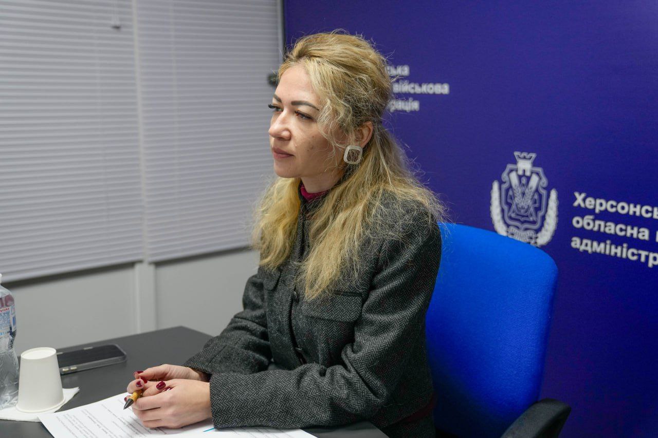 Жінка зі світлим волоссям сидить за робочим столом, тримаючи ручку над документами. Вона одягнена у сірий жакет; на фоні помітно синій банер із гербом та написом «Херсонська обласна військова адміністрація».
