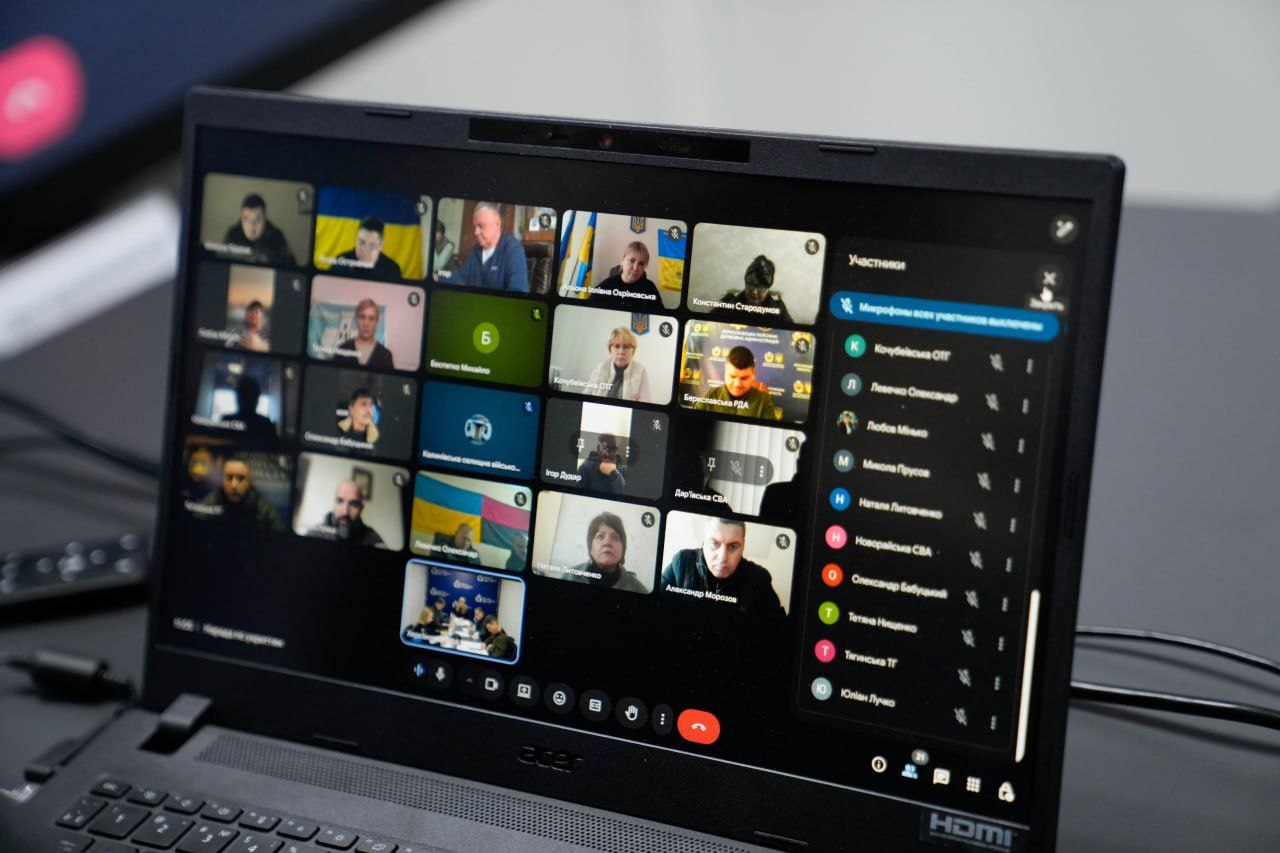 Екран відкритого ноутбука, на якому відображено вікно відеоконференції з багатьма учасниками у режимі сітки. Праворуч видно список присутніх; пристрій стоїть на робочому столі в офісному приміщенні.