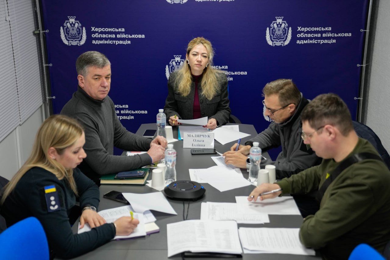П'ятеро людей сидять за столом під час робочої наради на фоні банера з офіційною символікою Херсонської обласної військової адміністрації. Учасники переглядають документи та роблять записи; на столі стоять пляшки з водою та паперові стаканчики.