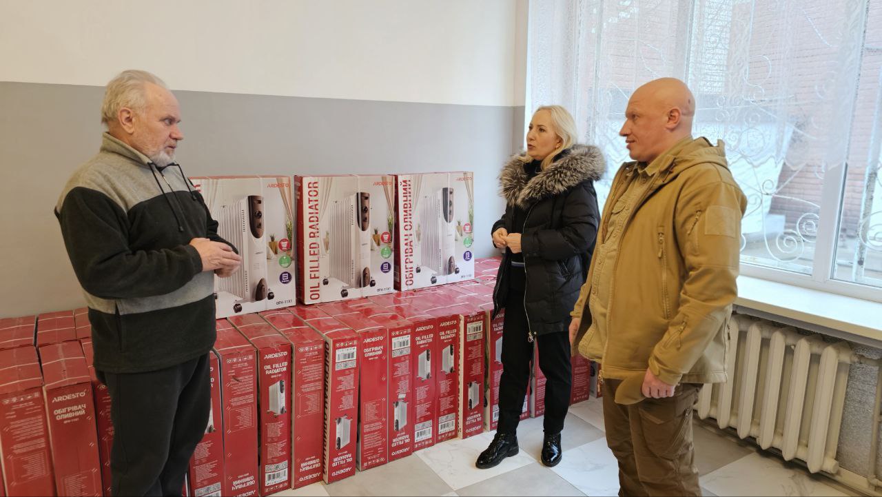 Троє людей спілкуються у приміщенні на фоні численних коробок з олійними радіаторами опалення. Чоловік та жінка у верхньому одязі розмовляють із чоловіком у темному светрі. Позаду них видно вікно з білою тюлю та чавунну батарею.