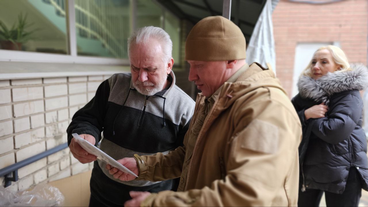 Двоє чоловіків на вулиці біля цегляної будівлі уважно вивчають документи. Один із них одягнений у сіро-чорну кофту, інший — у куртку гірчичного кольору та коричневу шапку. Позаду них стоїть жінка в чорній куртці з хутряним коміром.