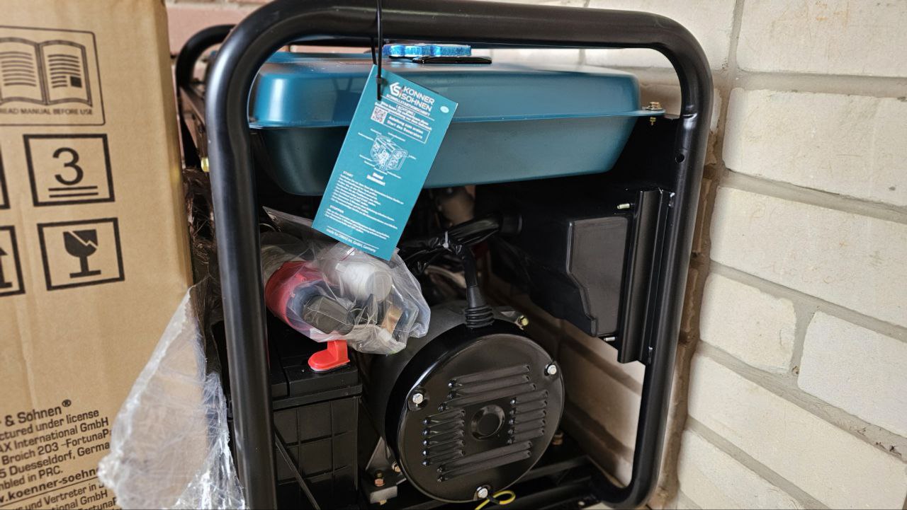 Дизельний генератор у металевому каркасі, що стоїть на вулиці біля цегляної стіни. Пристрій має бірюзовий паливний бак, до якого прикріплена інструкція у вигляді синьої картки. Поруч видно частину картонного пакування з технічним маркуванням виробника.