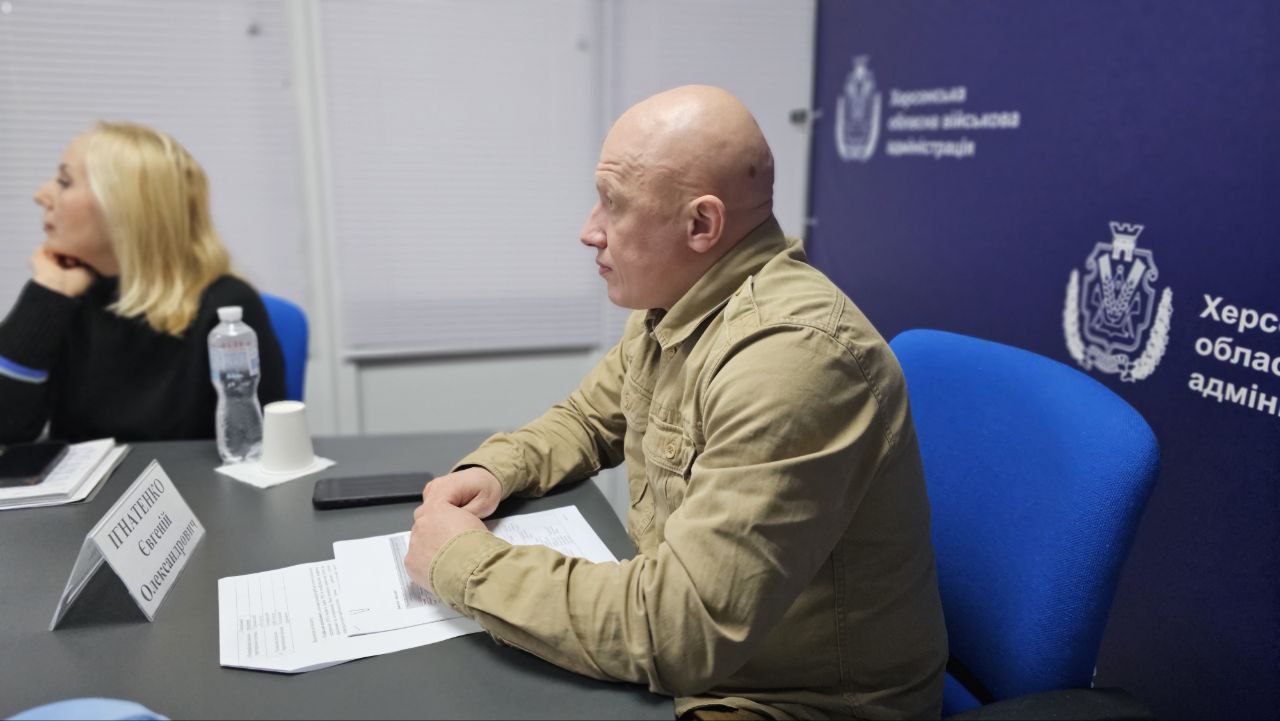 Чоловік у тактичній сорочці пісочного кольору сидить за столом в офіційному кабінеті. Перед ним лежать паперові документи, на столі стоїть табличка з іменем та пляшка води. На задньому плані — синій банер із логотипом Херсонської обласної військової адміністрації.