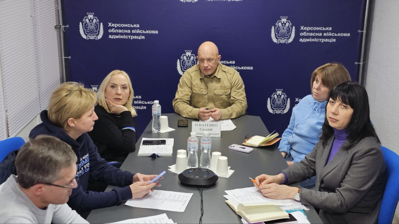 Робоча нарада в офіційному приміщенні з групою людей, які сидять за довгим столом. Учасники у цивільному та військовому одязі вивчають документи та ведуть обговорення. На задньому плані — синій банер з написом «Херсонська обласна військова адміністрація» та Державний Прапор України.