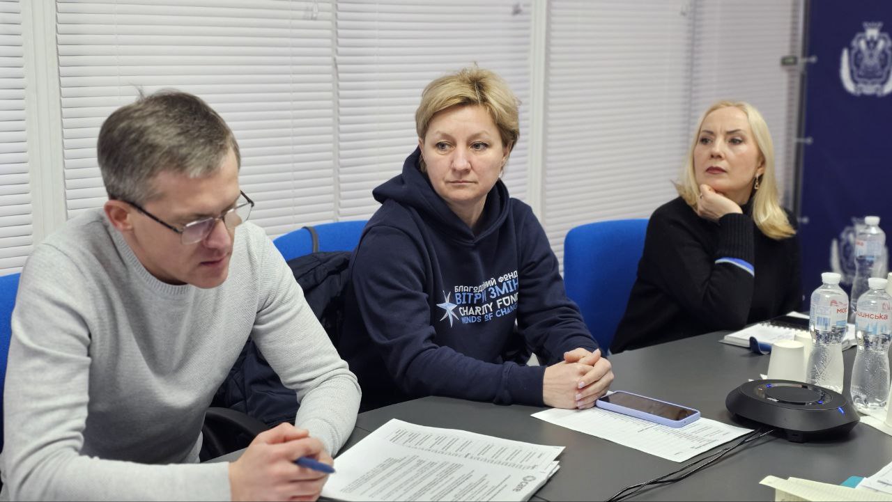 Троє учасників робочої зустрічі сидять за столом та уважно слухають доповідача. Чоловік на передньому плані вивчає документи, а жінка у синьому худі з логотипом благодійного фонду та ще одна учасниця дивляться в бік спікера.