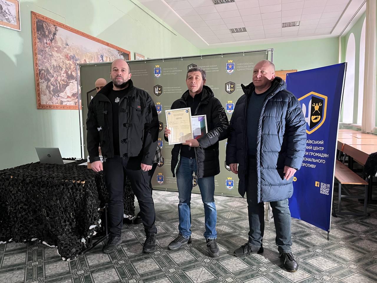 Троє чоловіків на фоні офіційного банера. У центрі — чоловік із коротким сивіючим волоссям у чорній куртці та джинсах, який тримає сертифікат. Поруч стоїть стіл, накритий маскувальною сіткою, з ноутбуком, а на стіні позаду — велика картина.