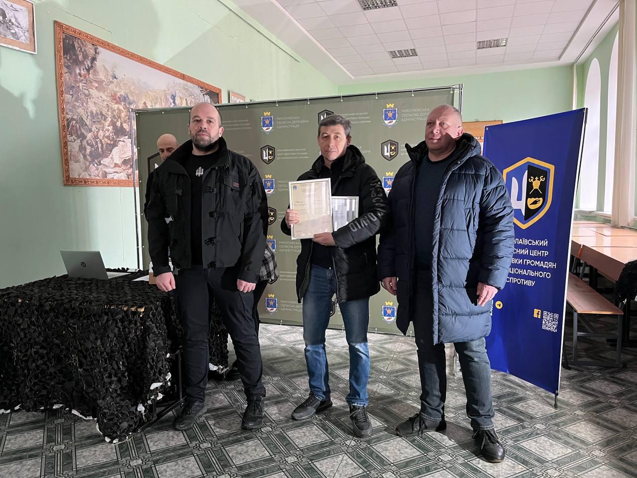 Троє чоловіків на фоні офіційного банера. У центрі — чоловік із коротким сивіючим волоссям у чорній куртці та джинсах, який тримає сертифікат. Поруч стоїть стіл, накритий маскувальною сіткою, з ноутбуком, а на стіні позаду — велика картина.