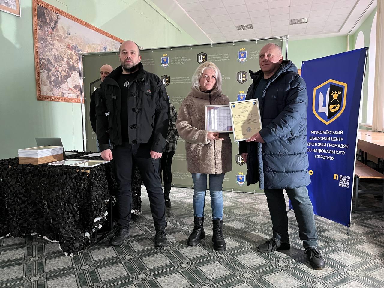 Троє людей на фоні офіційного банера Миколаївського обласного центру підготовки населення до національного спротиву. У центрі — жінка у світлій шубі та блакитних джинсах, яка тримає сертифікати. Поруч стоїть стіл, задрапірований маскувальною сіткою, а на стіні позаду видно велику картину.