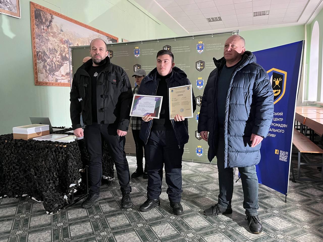 Троє чоловіків на фоні офіційного банера. У центрі — молодий чоловік у темному одязі, який тримає в руках два різні сертифікати. Справа від нього стоїть стіл, накритий маскувальною сіткою, з документами. На стіні ліворуч висить велике художнє полотно.