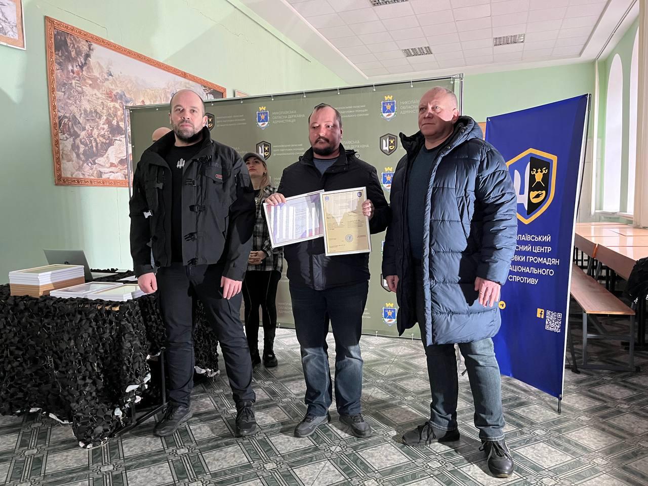 Чоловік із вусами та короткою борідкою, одягнений у темну куртку, стоїть у центрі та тримає дві рамки з нагородами. Він знаходиться між двома іншими чоловіками на фоні офіційного банера Миколаївської ОДА. Ліворуч на столі, накритому маскувальною сіткою, лежать стоси готових дипломів.