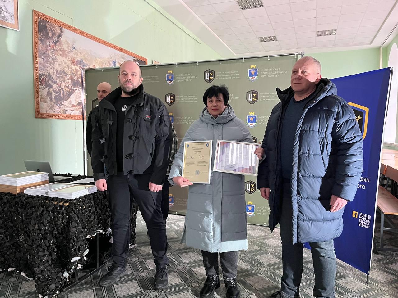 Жінка у світлому подовженому пальті стоїть у центрі групи, тримаючи дві рамки з нагородами. Вона знаходиться між двома чоловіками на фоні офіційного банера та картини з батальною сценою. Поруч на столі лежать стоси підготовлених сертифікатів.