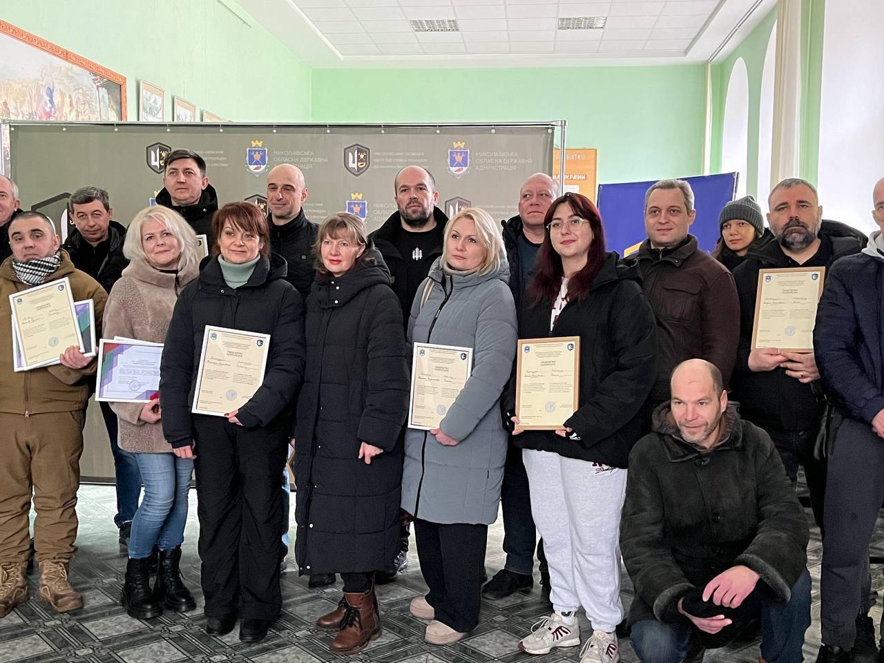 Група людей стоїть у приміщенні на фоні офіційного банера з гербами. Дехто з присутніх тримає в руках сертифікати або грамоти. Усі одягнені у верхній зимовий одяг. Атмосфера події виглядає офіційною та урочистою.