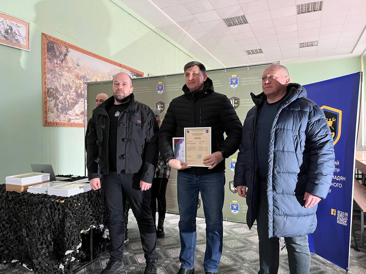 Троє чоловіків стоять у приміщенні на фоні банера з офіційною символікою Миколаївської області. Чоловік у центрі, одягнений у чорну стьобану куртку та сині джинси, тримає вертикальний сертифікат у світлій рамці. Ліворуч на столі, накритому маскувальною сіткою, лежать стоси дипломів, а на стіні позаду висить велика картина з батальною сценою.
