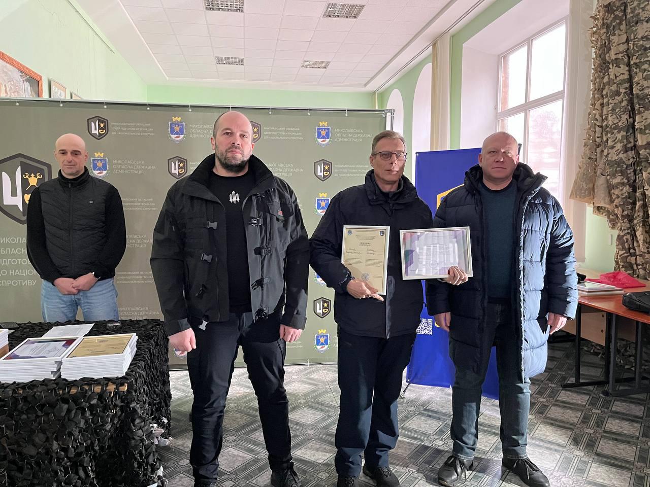 Група чоловіків стоїть у приміщенні на фоні офіційного банера Миколаївської ОДА. Один із них, чоловік в окулярах та темно-синій куртці, тримає перед собою вертикальний сертифікат у світлій рамці. Поруч на столі, накритому чорною маскувальною сіткою, лежать стоси нагородних документів.