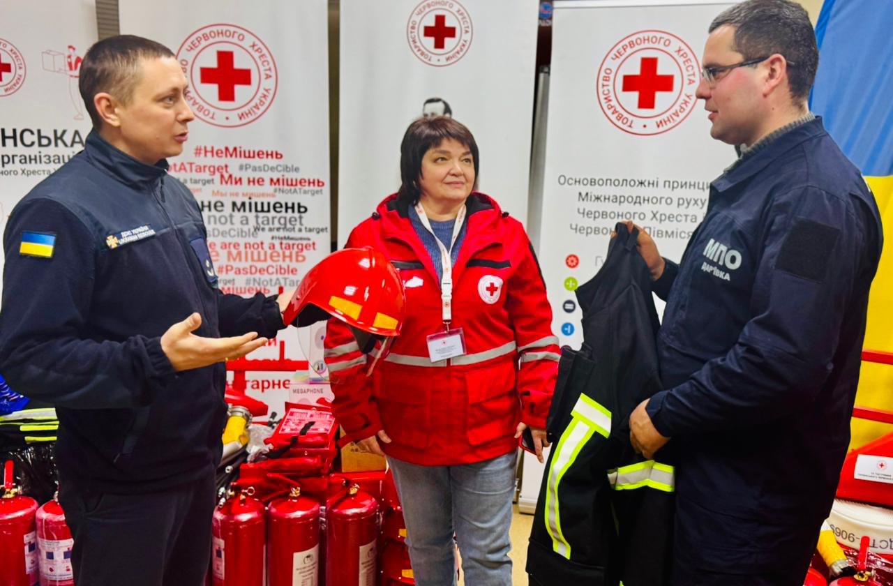 Три особи на фоні символіки Червоного Хреста обговорюють передачу рятувального спорядження. Чоловік у формі рятувальника жестикулює, жінка в червоній куртці тримає захисний шолом, а інший чоловік оглядає елемент спецодягу. На фоні видно вогнегасники та банери з написами «Ми не мішень».