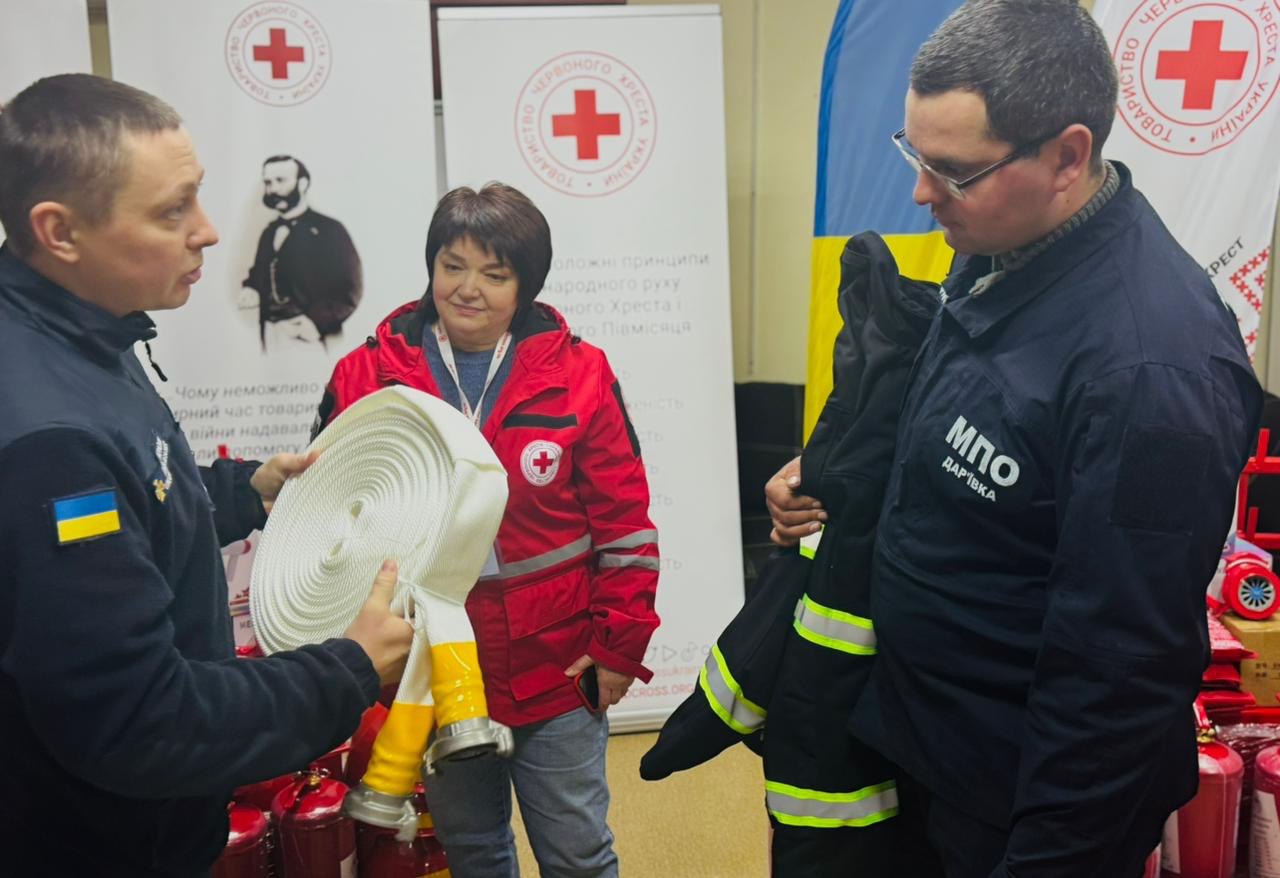Три особи на фоні банерів Червоного Хреста оглядають нове пожежне обладнання. Ліворуч чоловік у синій формі демонструє білий пожежний рукав. У центрі стоїть жінка в червоній куртці організації, а праворуч — чоловік у формі з написом «МПО», який тримає елемент захисного спецодягу. На передньому плані видно червоні вогнегасники.