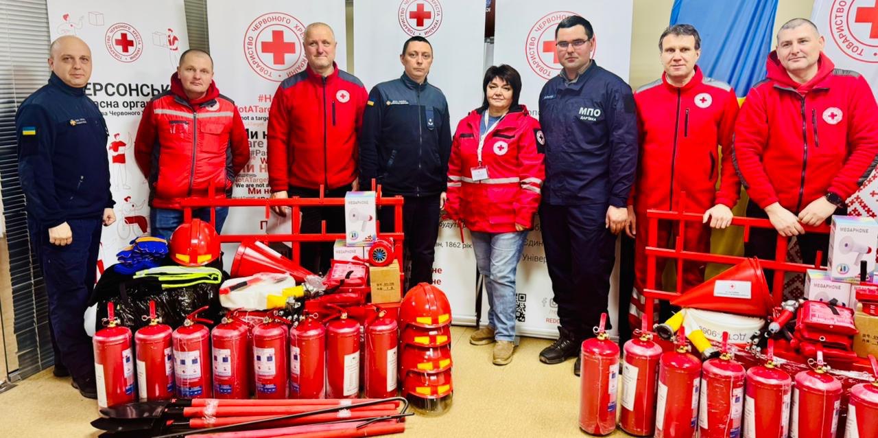 Група з семи чоловіків та однієї жінки стоїть у приміщенні на фоні банерів з емблемою Товариства Червоного Хреста України. Учасники одягнені в уніформу рятувальників та червоний фірмовий одяг організації. Перед ними на підлозі розкладено значну кількість пожежного та рятувального спорядження: численні вогнегасники, захисні шоломи, мегафони та пожежні інструменти.