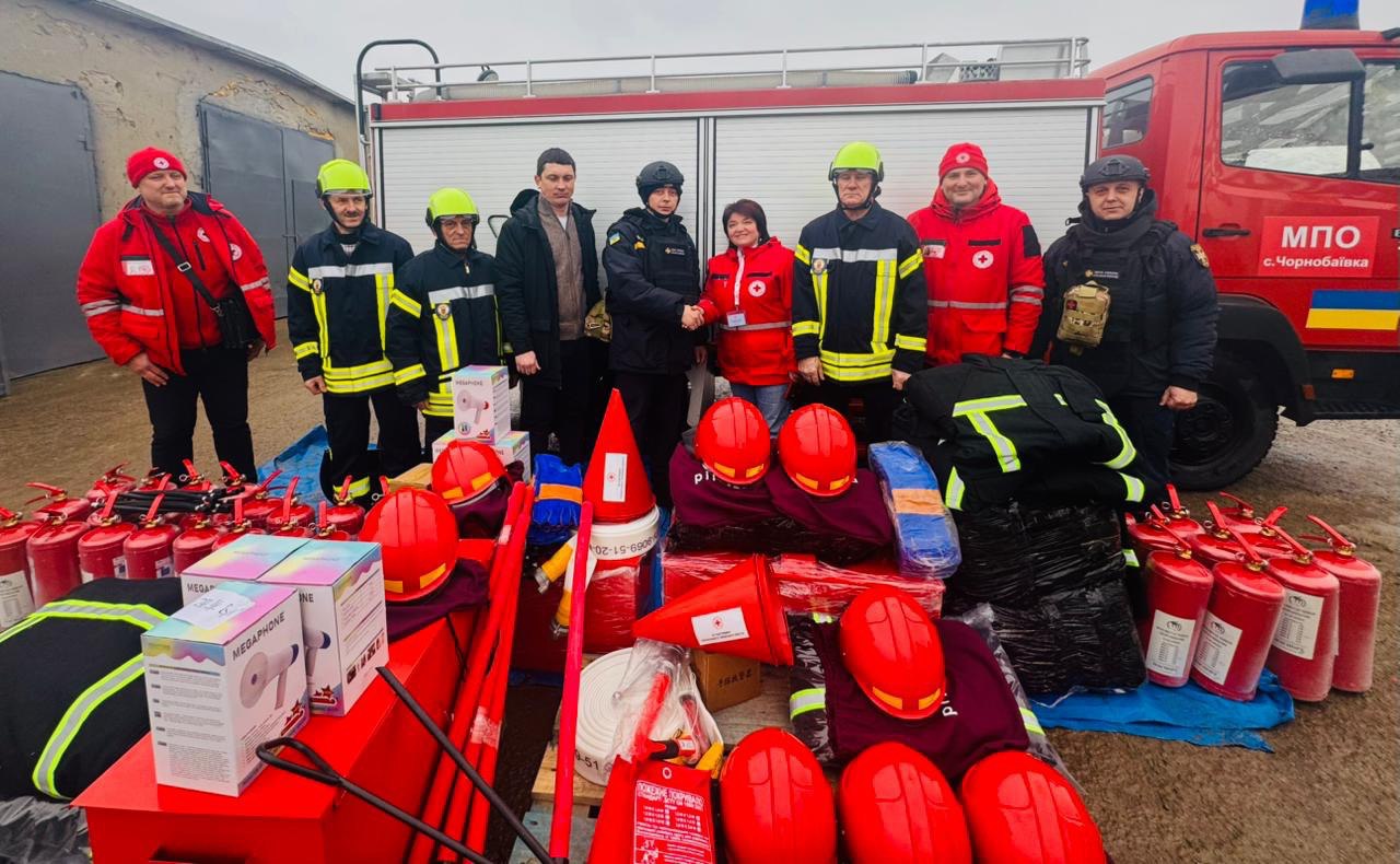 Група людей у формі рятувальників, пожежників та представників Червоного Хреста стоїть перед пожежною машиною та розкладеним на землі гуманітарним обладнанням. У центрі двоє осіб потискають одне одному руки. Попереду на столах та брезенті розкладено численні червоні захисні шоломи, вогнегасники, мегафони, пожежні рукави та інше спорядження.
