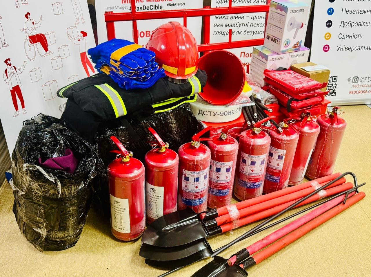 На підлозі розкладено комплект пожежного та рятувального інвентарю: вогнегасники, лопати, захисний шолом, рукавиці та спецодяг. Поруч стоять коробки з мегафонами на фоні банерів із символікою Червоного Хреста.