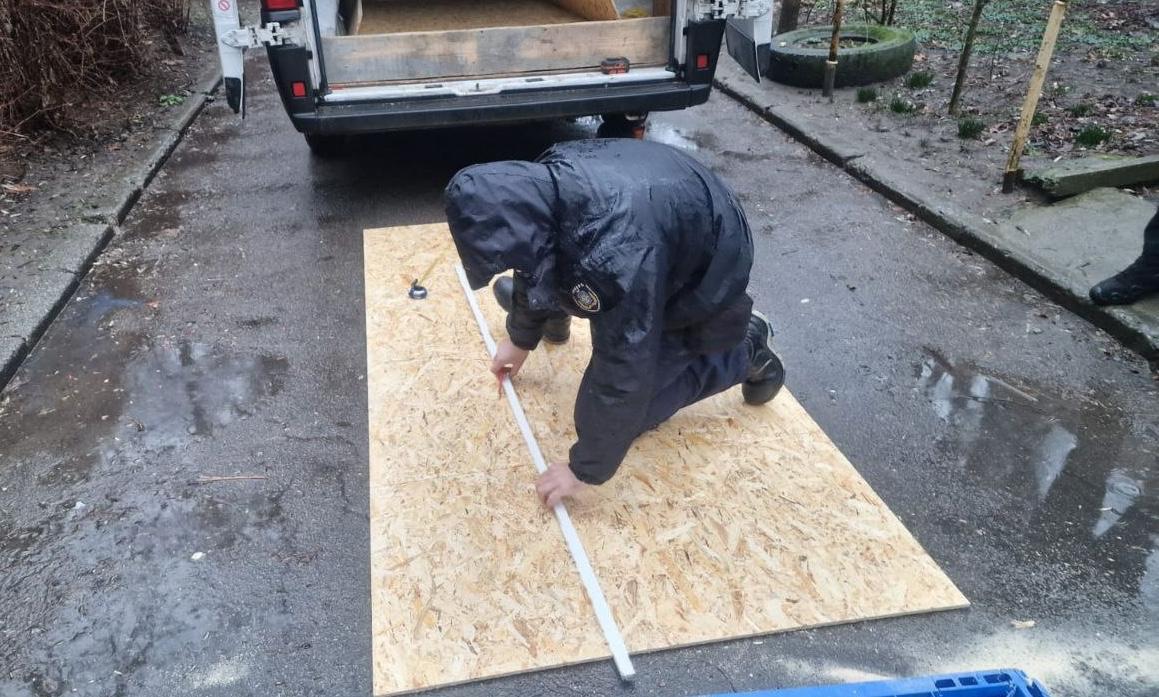 Спеціаліст у темному захисному одязі з капюшоном схилився над листом ОСБ-плити на асфальті, роблячи точні заміри та розмітку за допомогою довгої лінійки. Позаду видно відкритий багажник робочого автомобіля.